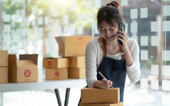 Happy young Asian business owner woman prepare parcel box and check online orders of product for deliver to customer on smartphone. Shopping Online concept..