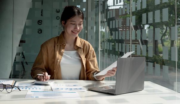 Young Asian businesswoman is happy to work at the modern office using laptop and calculator, working on project, checking financial documents.