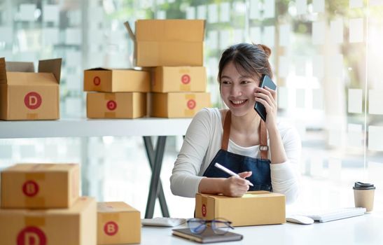 Small business entrepreneur SME freelance woman using phone call receive from customer checking product on stock at home office, online marketing packaging delivery box, SME e-commerce concept..