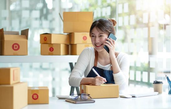 Small business entrepreneur SME freelance woman using phone call receive from customer checking product on stock at home office, online marketing packaging delivery box, SME e-commerce concept..