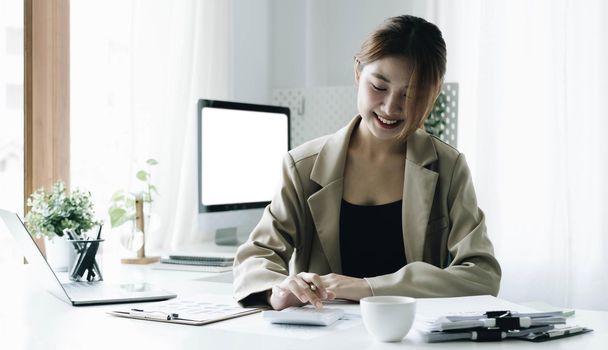 Smiling female accountant using calculator while sitting at office desk with digital tablet and financial documents..