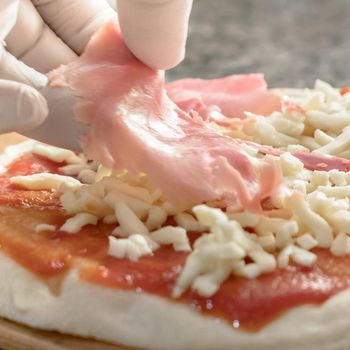 Hand of pizza chef with white gloves adds cooked ham on pizza.