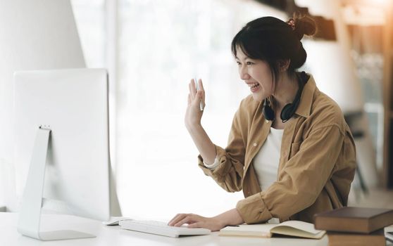 Head shot portrait smiling asian woman wearing headphones posing for photo at workplace, happy excited female wearing headset looking at laptop, sitting at desk with laptop, making video call.