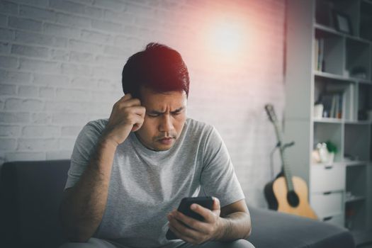Depression concept. Asian man sad and checking mobile phone sitting on the sofa in the living room at home, Asian man serious and worry while using mobile phone.