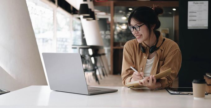 Happy asian businesswoman wearing headset make notes talk by conference call doing video chat on laptop in office, smiling chinese support service agent write information consult customer.