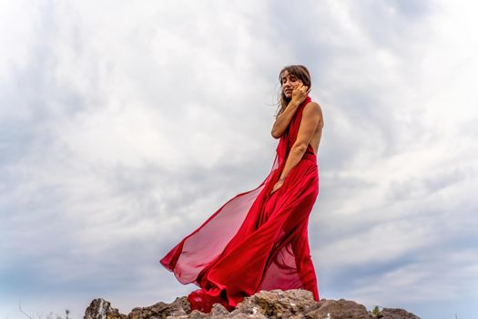 A woman in a red dress stands above a stormy sky, her dress fluttering, the fabric flying in the wind