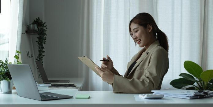 Attractive young woman sitting and her workplace and checking information on clipboard..