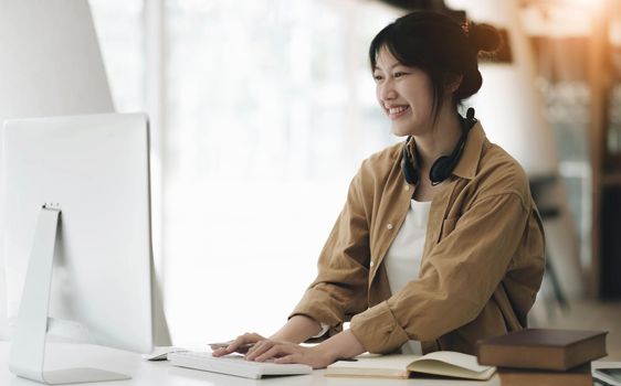Head shot portrait smiling asian woman wearing headphones posing for photo at workplace, happy excited female wearing headset looking at laptop, sitting at desk with laptop, making video call.