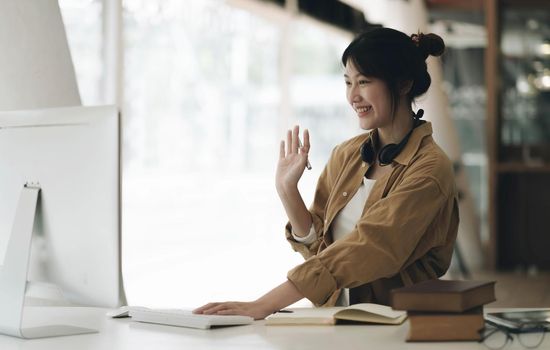 Asian woman wearing headphones on her neck using laptop and greeting friends on video call at home