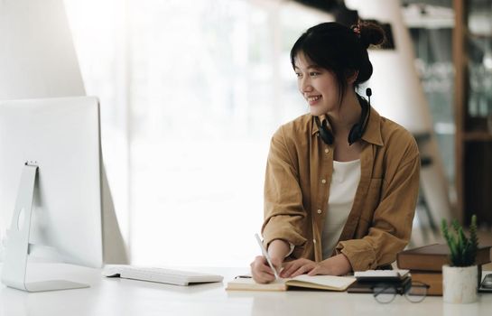 Asian woman wearing headphones using laptop and taking notes on homework that her teacher teaches at home