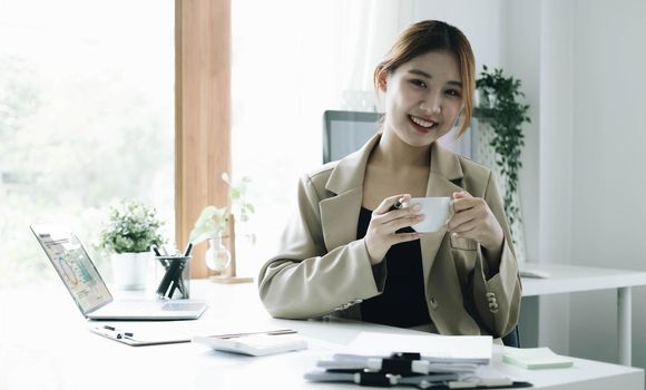 Smiling Asian businesswoman holding a coffee mug and laptop at the office. Looking at the camera..