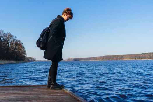 girl in a black coat stands at the edge of the pier and looks into the water of the lake