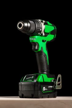 a cordless screwdriver stands on a wooden table on a black background. Cordless drill with lithium-ion battery in green. Professional tool for drilling holes and driving screws