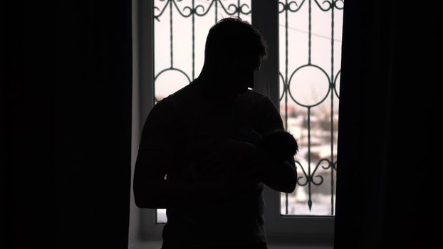 A young dad holds a child in his arms against the background of the window. The light from the window illuminates the image of a man with a newborn child. 4k