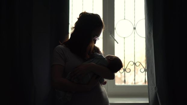 A young mother holds a child in her arms against the background of a window. The light from the window illuminates the image of a woman with a newborn baby. 4k