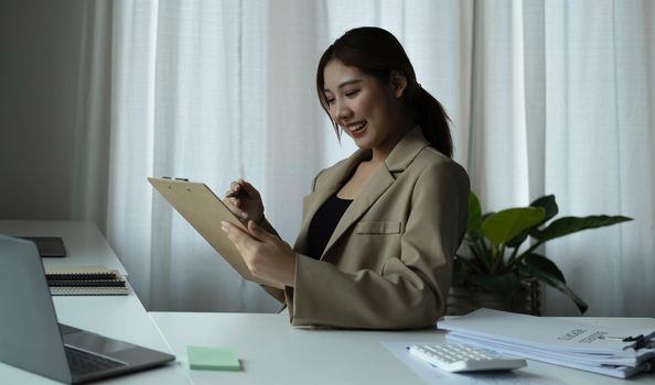 Attractive young woman sitting and her workplace and checking information on clipboard..