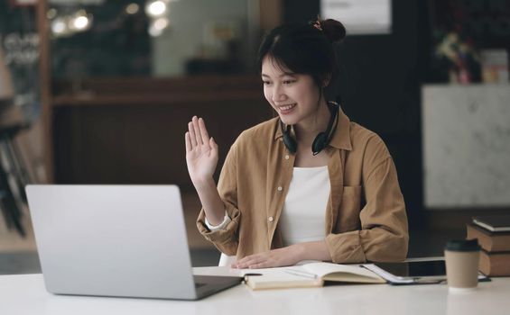 Asian woman wearing headphones on her neck using laptop and greeting friends on video call at home