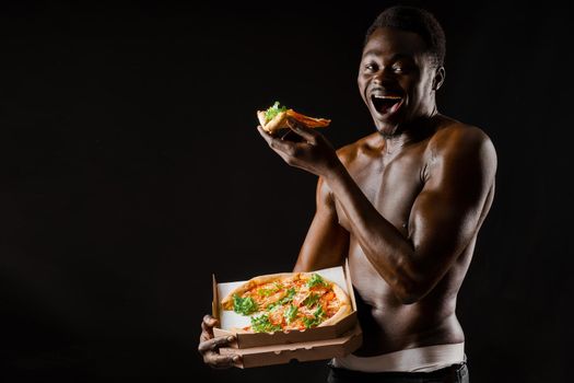 Black handsome naked man eats a pizza. Safety food delivery for people who stays at home. Sexy african man shows 2 pieces of pizza in his hands.
