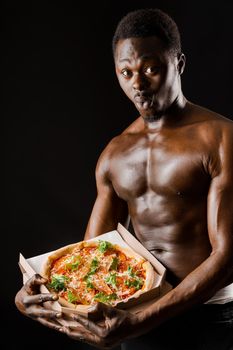 Surprised black fit man with pizza in hands. Tasty food delivery. African with athletic body shows a pizza. Sexy man with tasty pizza from restaurant