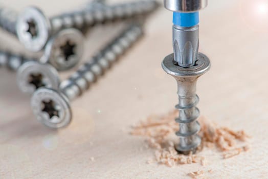 Screw the screw into the board. Furniture production, a screwdriver twists a self-tapping screw into a board. Several silver screws lie on the desktop.