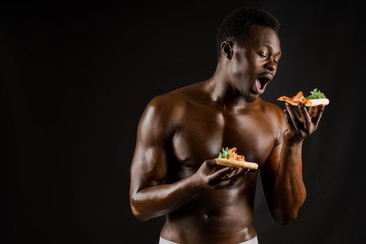 Black handsome naked man eats a pizza. Safety food delivery for people who stays at home. Sexy african man shows 2 pieces of pizza in his hands