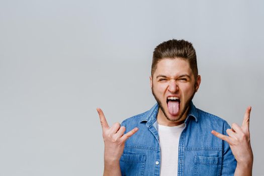 Confident bearded man shows tongue and makes rock n roll. Advertise for party. Young handsome man stants over white background and shows cool.