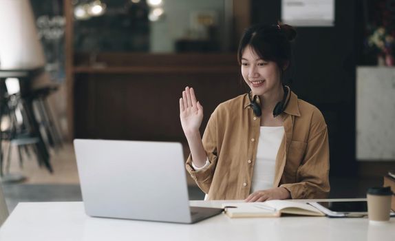 Asian woman wearing headphones on her neck using laptop and greeting friends on video call at home