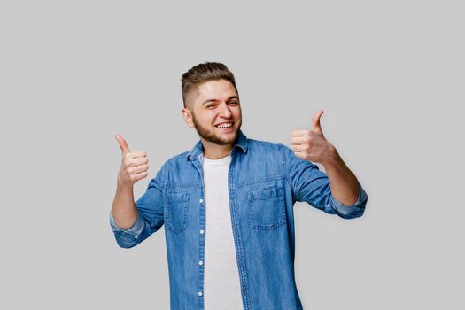 Confident bearded man wears denim shirt and shows thumbs up. Advertise for party. Young handsome man stants over white background and shows cool.