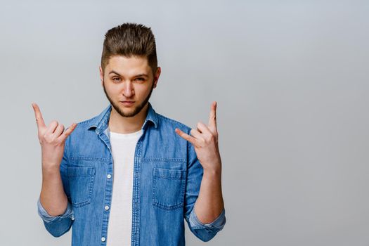 Confident bearded man wears jeans shirt and makes rock n roll. Advertise for party. Young handsome man stants over white background and shows cool.