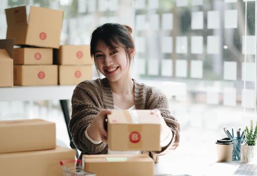 Happy young asian woman startup small business freelance holding parcel box and computer laptop and sitting on chair, Online marketing packing box delivery concept.