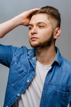 Young handsome man stands over white background and touches his hair after visiting barbershop. Confident man wears casual jeans shirt