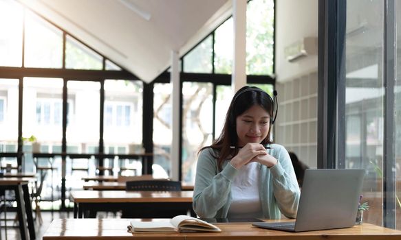 Smiling girl student wear wireless headphone study online with skype teacher, happy young woman learn language listen lecture watch webinar write notes look at laptop sit in cafe, distant education.