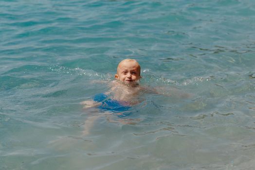 Child learning to swim in the open sea of tropical resort. Kids learn swimming. Exercise and training for young children. Little boy with colorful float board in sport club. Swimming baby or toddler. Happy child boy swims in sea in swimming circle with splash. Blue sky and water. Swimming training. Fun joy activities on vacation in the beach. Childhood moments lifestyle. Freedom careless. boy swim in the sea.