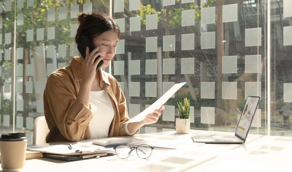 Charming Asian woman with a smile standing holding papers and mobile phone at the office..