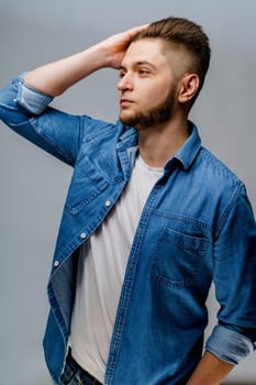 Young handsome man stands over white background and touches his hair after visiting barbershop. Confident man wears casual jeans shirt