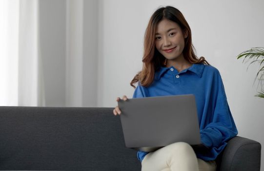 Happy smiling Asian woman working on laptop computer while sitting on sofa and looking at camera.