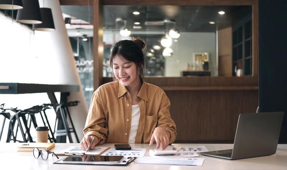 Happy young asian businesswoman sitting on her workplace and using calculator in the office. Young woman working at laptop in the office..