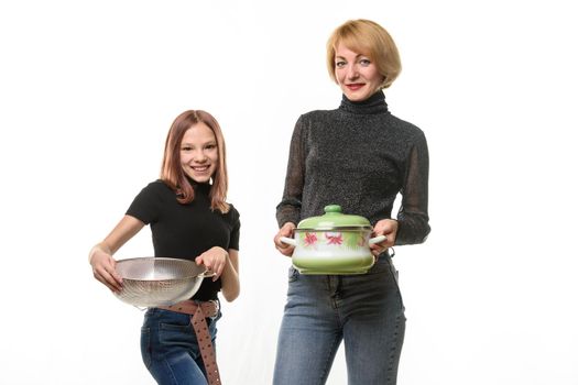 Mom stands with a saucepan, daughter stands with a sieve, both smile and look into the frame