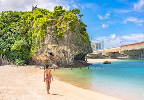 Young woman in swimsuit from behind on the sandy beach Naminoue topped by a huge rock with a Shinto Shrine at the top of a cliff and a highway passing in Naha City in Okinawa Prefecture, Japan.