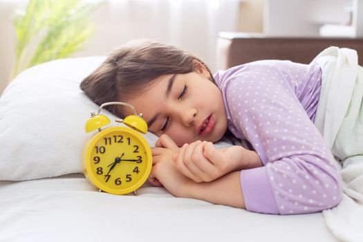 A sleeping brunette girl in purple polka dot pajamas lie on a white pillow, on a bed, in the morning light, a yellow alarm clock stands nearby. copy space
