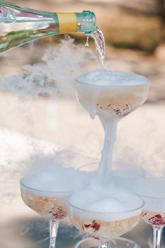 The waiter pours champagne into crystal glasses with dry ice and white smoke close up. Sparkling wine pouring in wine glass at summer day.