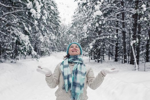 Snow is falling from above on a happy girl in a white sweater, blue hat and scarf, stands in the winter forest