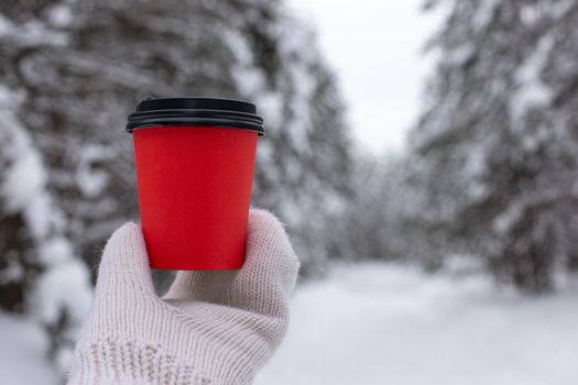 Red paper cup of coffee in hand in a white knitted mittens, against the backdrop of a beautiful winter forest. Copy space. Close up