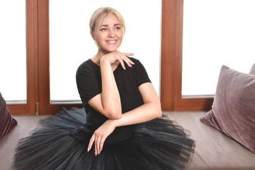 a young, smiling ballerina in a black tutu sits in front of the window with her chin tucked in