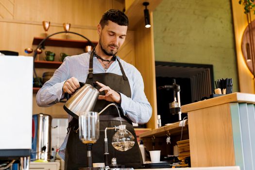 Handsome bearded barista pours hot boiling water from kattle to syphon device for coffee brewing in cafe. Syphon alternative method of making coffee.
