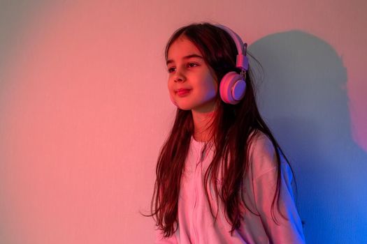 A happy little girl in pink headphones, with loose dark hair, stands against the wall in neon pink-blue light