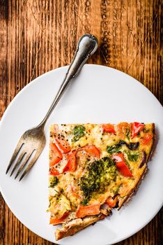 Slice of vegetable frittata with broccoli, red bell pepper and red onion on white plate. View from above