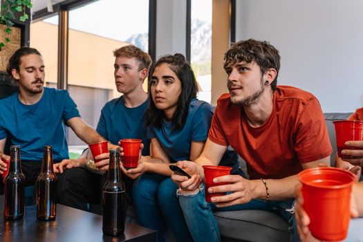 friends together watching television in concentration. watching a movie. watching football on television. sitting on the couch in the living room, natural light. table with drink and food.