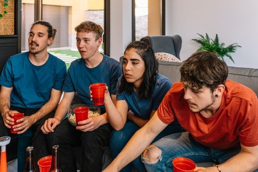 friends enjoying together a sports match broadcast on television. watching a movie. eating and drinking. sitting on the sofa in the living room, natural light.