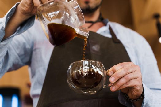 Close-up pouring coffee in double glass cup in cafe by handsome bearded barista. Coffee brewing syphon and aeropress alternative methods